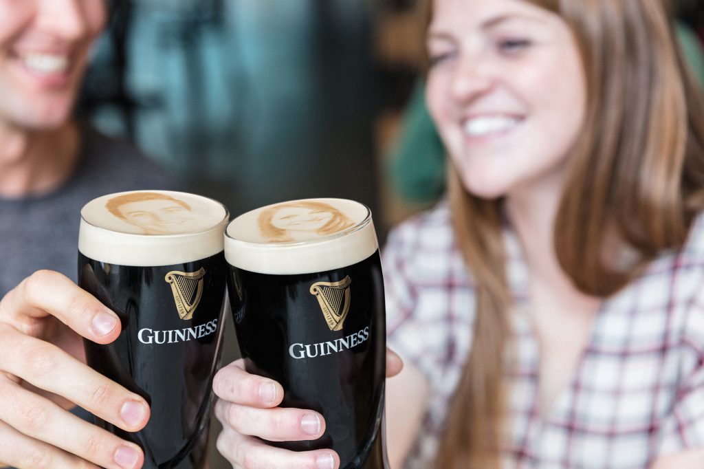 Two people holding pints of Guinness with images of themselves imprinted onto the heads of the pints.