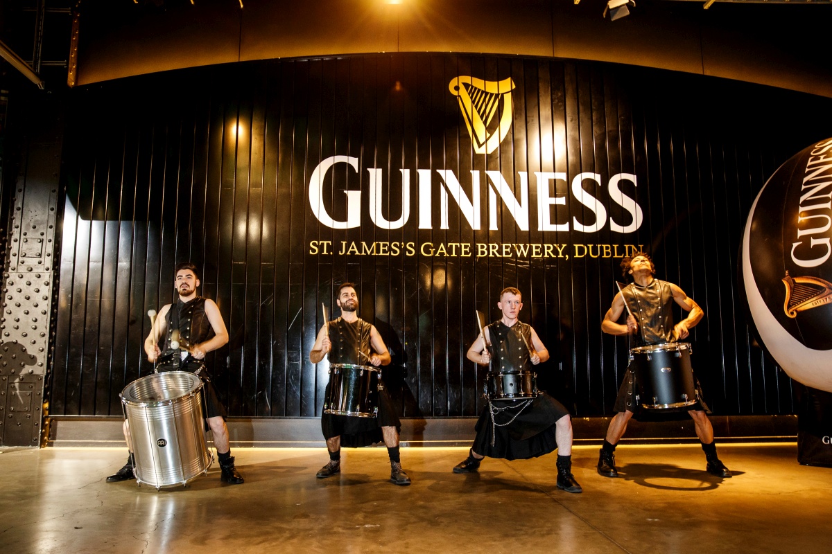 A group of men in leather waistcoats and kilts banging big drums in front of the Guinness St James's Gate Brewery